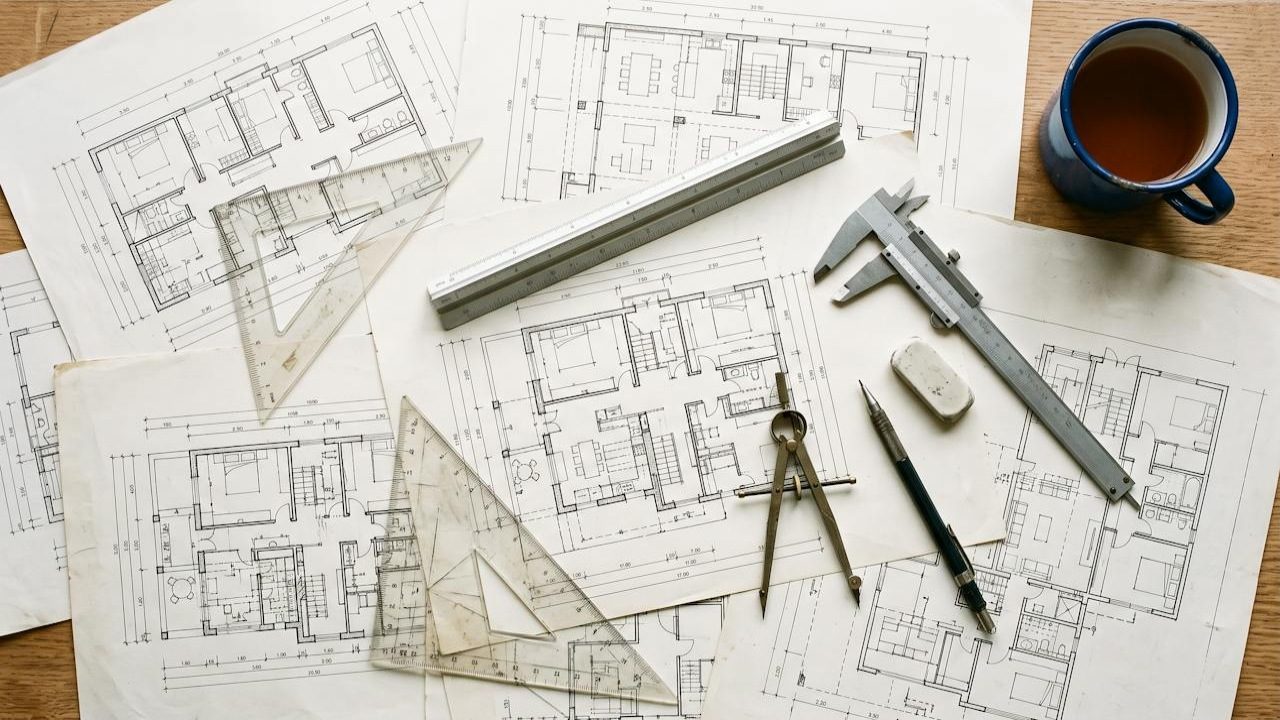 A drafting board covered with overlapping A3 architectural plans, a scale rule, set squares, calipers and a pencil.